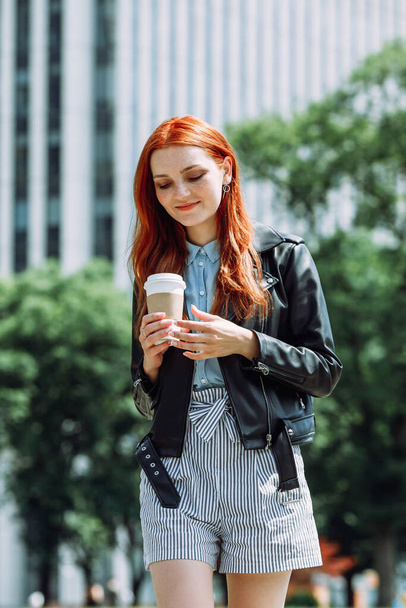 Happy young trendy red hair woman wearing black leather jacket and shorts, drinking take away coffee and walking in an urban city. City walk lifestyle concept. - Photo, image