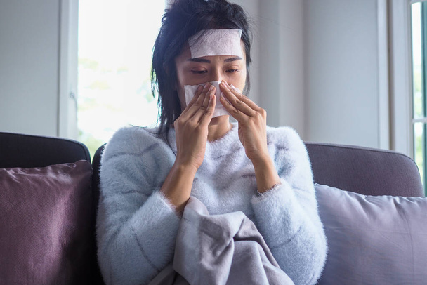 Sick woman sitting on the sofa in the house, having high fever on the forehead with a gel pad to reduce body temperature, sore throat, runny nose and coughing. - Photo, Image