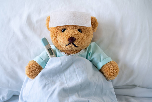 El oso enfermo lleva ropa de paciente. acostado en la cama con fiebre, un gel reductor de fiebre en la frente y en el templo de temperatura, lo puso debajo de la axila
 - Foto, Imagen