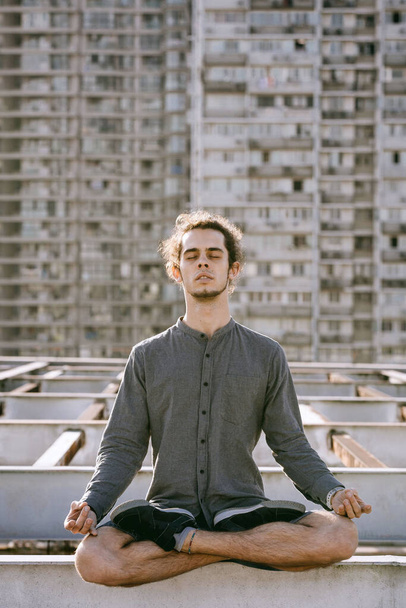 Meditating young european man sitting with eyes closed in lotus pose on blurry urban residential building. Yoga concept - Фото, изображение