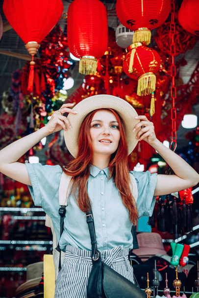 young caucasian red hair woman tries on a hat while shopping at a store that sells wicker hats in Asian market in China - Foto, Imagem