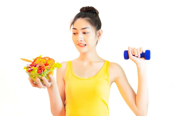 Jonge mooie aziatische vrouw droeg geel ondershirt, Holding een salade glazen beker geïsoleerd op witte achtergrond met dummbell - Foto, afbeelding