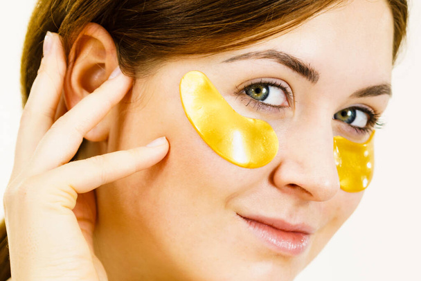 Young woman applying golden collagen patches under eyes. Mask removing wrinkles and dark circles. Girl taking care of delicate skin around eye. Beauty treatment. - Φωτογραφία, εικόνα