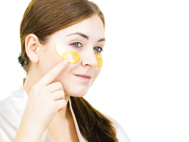 Young woman applying golden collagen patches under eyes, on white. Mask removing wrinkles and dark circles. Girl taking care of delicate skin around eye. Beauty treatment. - Photo, Image