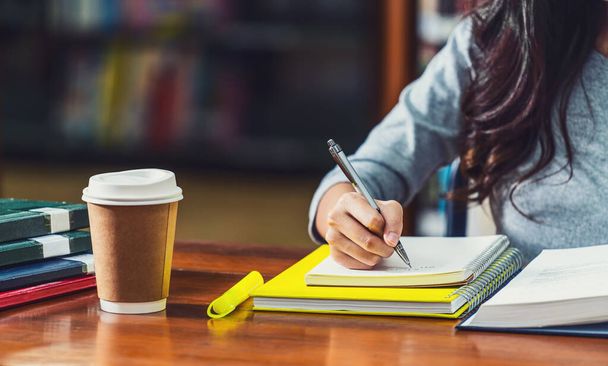 Nahaufnahme Asiatische junge Studentin schreibt Hausaufgaben in der Bibliothek der Universität oder Kollegen mit verschiedenen Büchern und Schreibwaren mit Kaffeetasse auf Holztisch über dem Bücherregalhintergrund, Zurück zur Schule - Foto, Bild