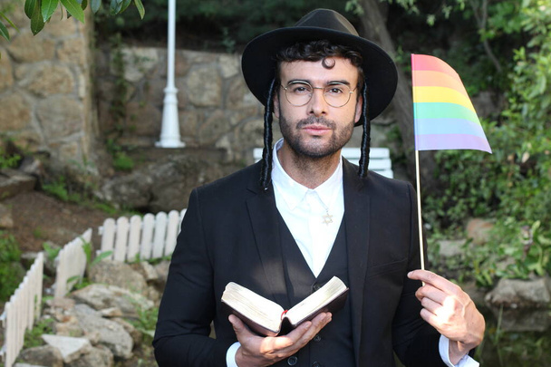 close-up portret van knappe jonge rabbijn met traditionele hoed met Tanakh lgbtq vlag op natuurlijke achtergrond - Foto, afbeelding