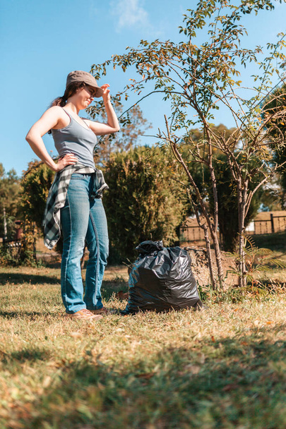 El concepto de contaminación ambiental y el día de la Tierra. Una voluntaria saca la basura en el parque, cerca de los pies hay una bolsa de basura negra. Orientación vertical
. - Foto, imagen