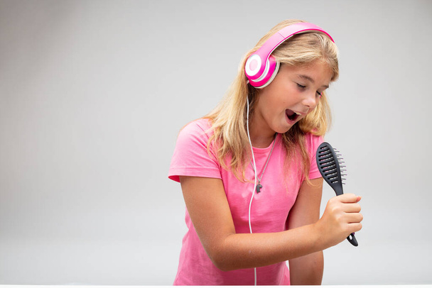 Молода дівчина весело співає в щітку для мікрофона, коли слухає свою музику на рожевих стерео навушниках на білому тлі
 - Фото, зображення
