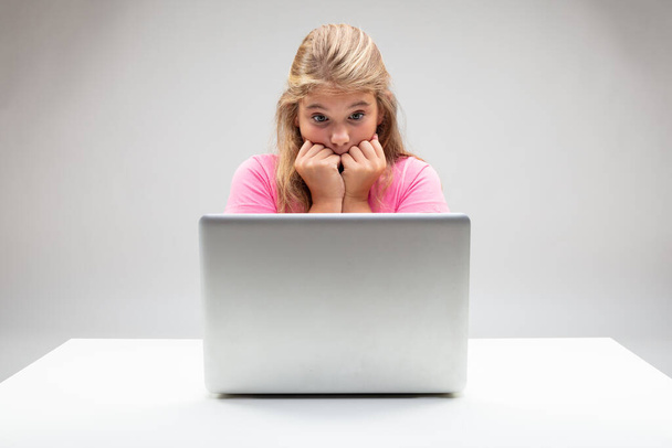 Chica joven mirando horrorizada a su computadora portátil con las manos en la boca y los ojos abiertos sentados en una mesa blanca sobre un fondo de estudio
 - Foto, Imagen