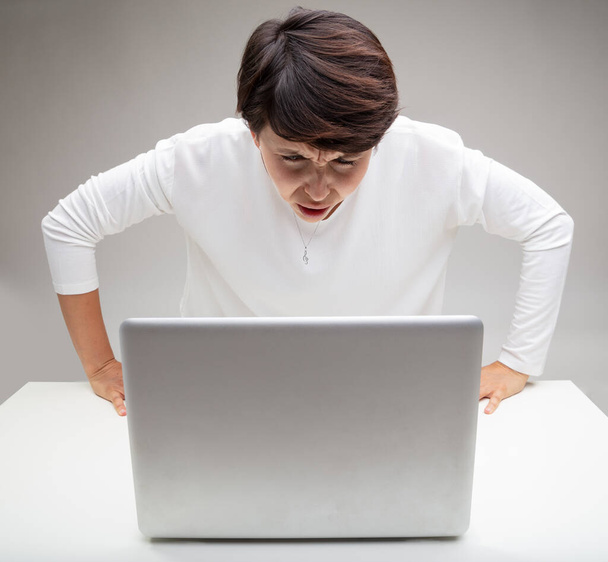 Stupefied nainen tuijottaa hänen kannettava tietokone kauhistuttava ilme epäuskon, kun hän nousee jaloilleen nojaten pöydälle lähikuva yli harmaa tausta - Valokuva, kuva