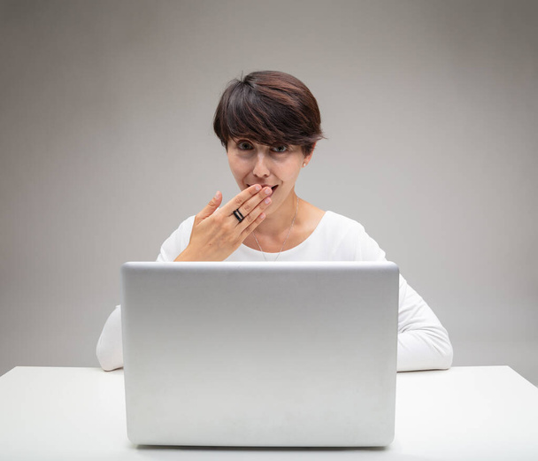Γεμάτη γυναίκα που καλύπτει το στόμα της καθώς γελάει με τον εαυτό της στη διασκέδαση καθισμένη πίσω από έναν φορητό υπολογιστή σε γκρι φόντο - Φωτογραφία, εικόνα