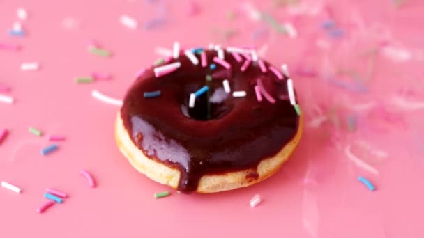 Donut brillante con acristalamiento de chocolate. Salpicaduras de colores cayendo sobre la rosquilla sobre fondo rosa
 - Imágenes, Vídeo