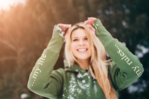 Blue-eyed blonde in a green hoody in the winter in the forest - focus on the sleeves with the inscription "my life - my rules" - Фото, зображення