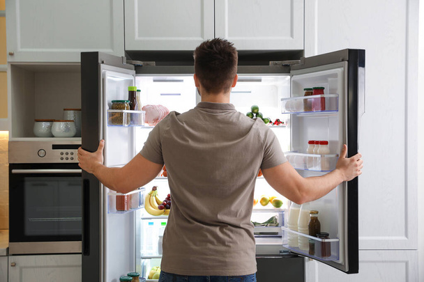 Young man opening refrigerator in kitchen, back view - Φωτογραφία, εικόνα
