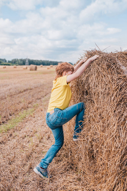 English red-haired boy climbs a large haystack - summer vacation in the village - Foto, immagini