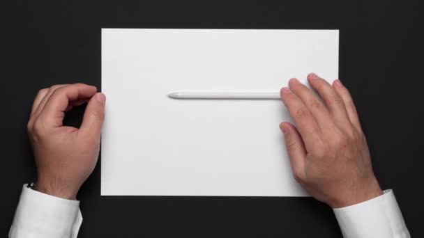 bovenaanzicht van een blanco vel papier en de handen van een zakenman op een zwarte tafel, wit shirt en polshorloge - Video