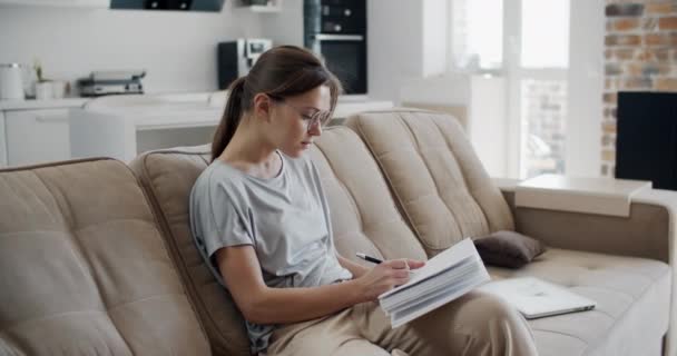 Young brunette woman works at home. A young woman sits on a sofa and makes notes in a notebook. Work at home during isolation. Female portrait. - Materiaali, video