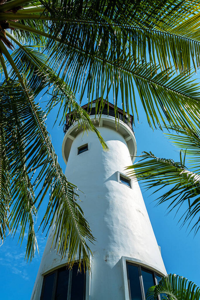 The white monolite structure of the lighthouse rises overover a palms tree. Thailand, Bangok. The massive tower of an old landmark that above the trees with clear blue sky on background. - Fotó, kép