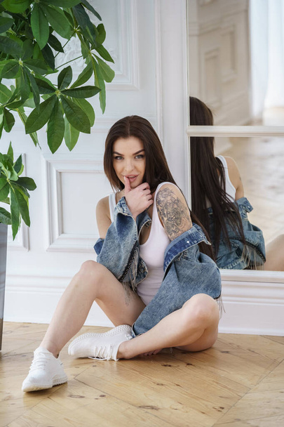 Attractive brunette woman wearing white lingerie, white sneakers and jeans jacket posing next to flowers pot at luxury apartments  - Fotoğraf, Görsel