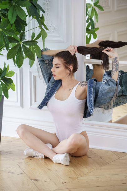 Attractive brunette woman wearing white lingerie, white sneakers and jeans jacket posing next to flowers pot at luxury apartments  - Φωτογραφία, εικόνα