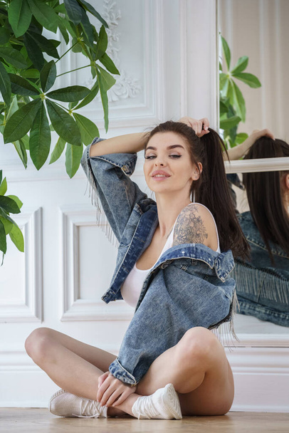 Attractive brunette woman wearing white lingerie, white sneakers and jeans jacket posing next to flowers pot at luxury apartments  - Фото, изображение