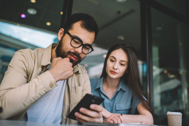 From below serious adult distrustful woman focusing on screen while happy ethnic man in glasses and casual clothes interacting with smartphone sitting at table with takeaway hot drink in modern cafe - Foto, imagen