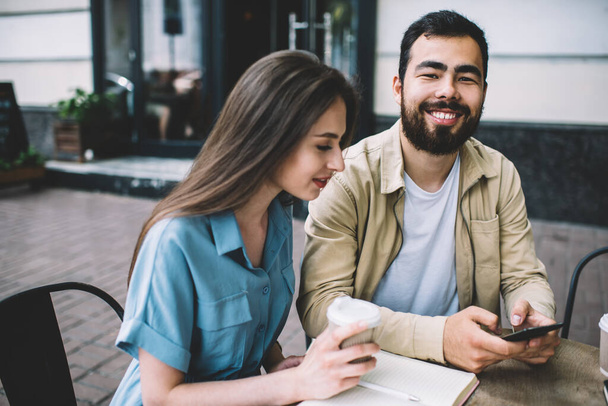 Happy bearded guy in casual clothes smiling and browsing smartphone while sitting near female colleague with takeaway beverage during break in remote job - Photo, image