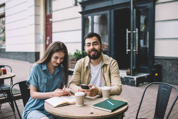Ethnic bearded man and young woman smiling and browsing smartphones while sitting at restaurant table during break in remote job on city street - Photo, image