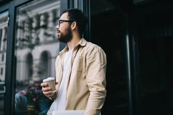 Confident modern busy bearded male in beige jacket and eyeglasses with laptop in hand holding cup of takeaway coffee while leaving coffee shop  - Valokuva, kuva