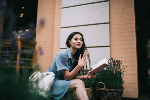 Stylish young woman with smartphone and planner looking away while sitting near baskets with flowers and studying on city street - Fotoğraf, Görsel