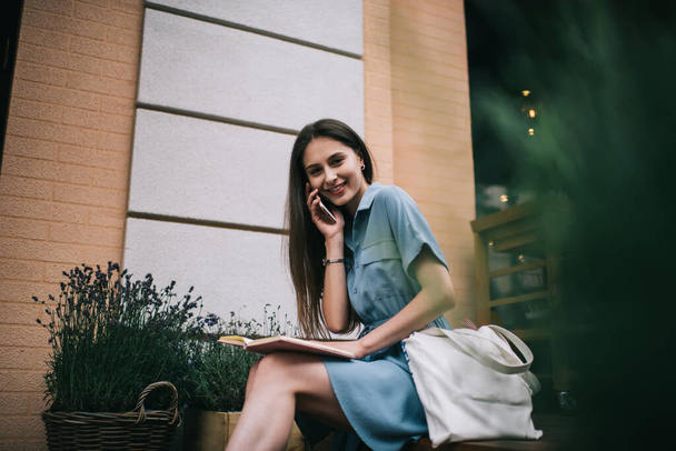 Delighted young woman with open planner smiling and looking at camera while sitting on bench outside building and talking on smartphone - Foto, Bild