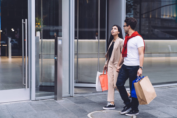 Side view pleased couple in trendy clothes shopping with paper bags full of purchases in hands and walking at street near with panoramic windows  - 写真・画像