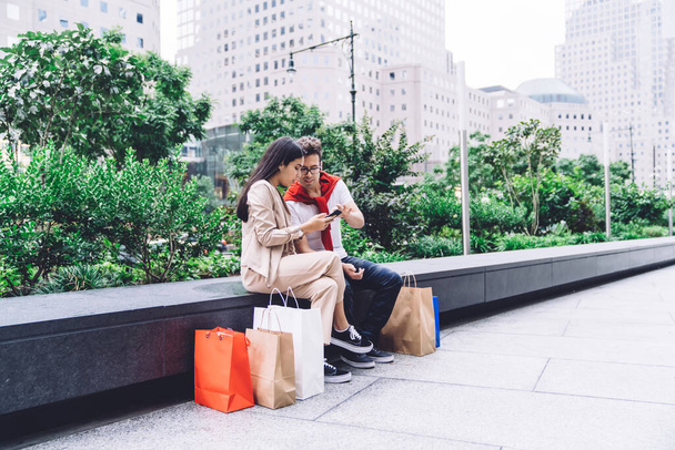 Trendy thoughtful modern casual multiracial young man and woman with shopping bags browsing on smartphone while sitting on parapet on city street  in daylight - Foto, Imagen