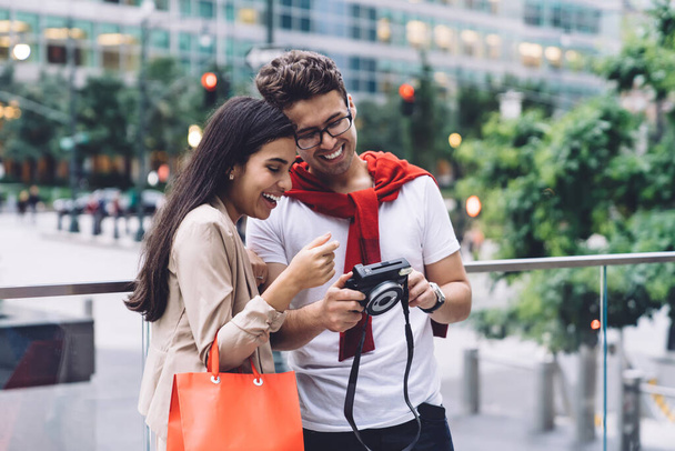 Smiling multiracial loving couple with bright shopping bag bonding on megapolis street while viewing photo camera in daylight on blurred background - Foto, immagini