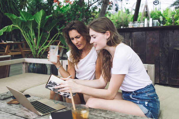 Focused young women with refreshing beverage reading notebook and using laptop while sitting in outdoor cafe in garden and studying together - Photo, Image