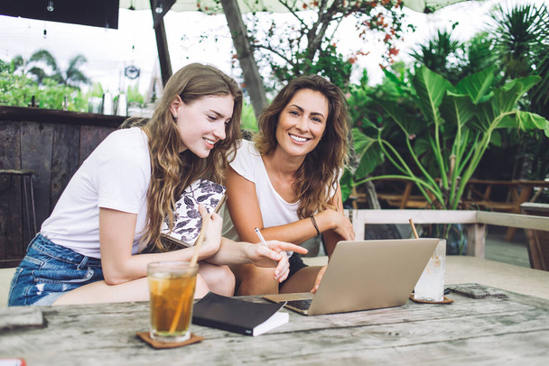 Fröhliche junge Damen lächeln und erledigen Hausaufgaben am Laptop, während sie am Tisch im Café im tropischen Garten sitzen - Foto, Bild