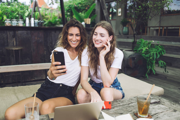 Optimistic female friends in stylish outfits smiling and using smartphone to take selfie while sitting on couch in outdoor restaurant - Fotó, kép