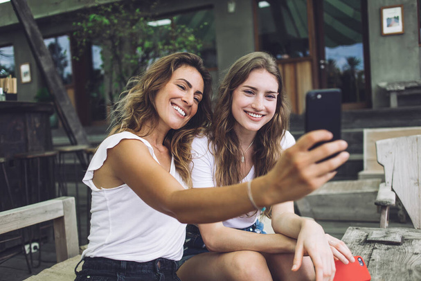 Cheerful trendy female smiling and using smartphone to take selfie with young friend while sitting on couch in cozy outdoor cafe - Фото, изображение