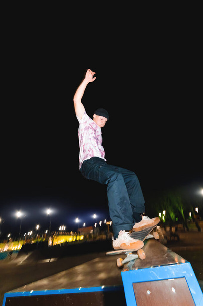 A young skater does the trick of sliding at night in a skatepark amid the burning lights of lighting poles. The concept of night leisure and youth culture at night - Foto, afbeelding