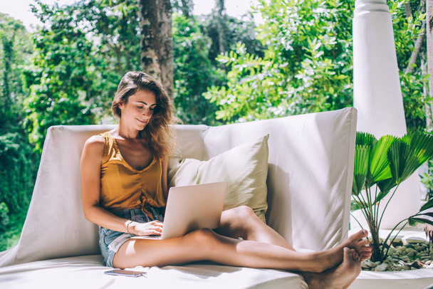 Güneşli bir günde bahçede beyaz bir kanepede otururken günlük giysiler içindeki güzel esmer kadın dizüstü bilgisayarda çalışıyor. - Fotoğraf, Görsel