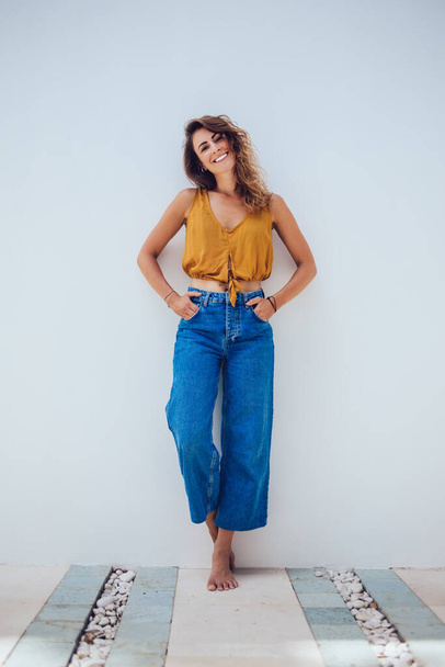 Optimistic content bronzed barefoot woman in casual clothing posing by wall with hands in pockets on white background and looking at camera - Фото, зображення
