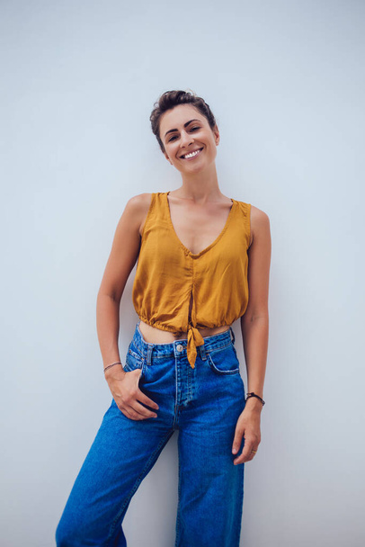 Stylish woman in jeans and orange shirt with hands in pocket smiling and looking at camera relaxed leaning on wall - Foto, Imagem