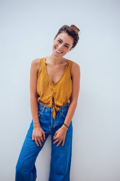 Joyful stylish laughing young lady wearing trendy orange top and blue jeans leaning on wall and laughing while posing in studio on white background - Foto, afbeelding