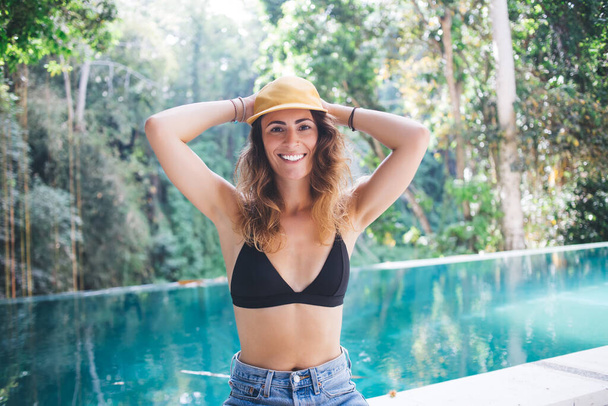 Beautiful smiling casual pleased stylish casual young woman in shorts and cap relaxing on poolside in peaceful yard on summer day - Zdjęcie, obraz
