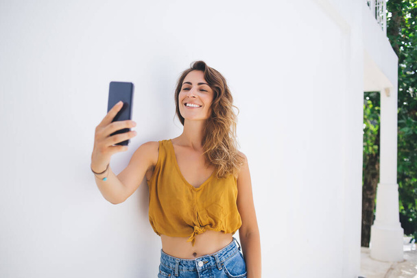Carefree pretty woman with bright smile in crop top leaning on whitewashed wall while keeping head up and talking selfie with smartphone - Photo, image