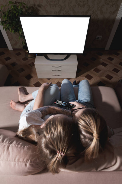 Aantrekkelijk jong langharig echtpaar knuffelend op een bank thuis in isolatie tv aan het kijken. Het concept van tijd doorbrengen met geliefden tijdens een pandemie. - Foto, afbeelding