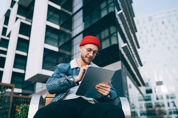 From below delighted fashionable bearded young man in glasses and red hat and denim jacket sitting on wooden bench and tapping on tablet on blurred urban background - Foto, Imagem