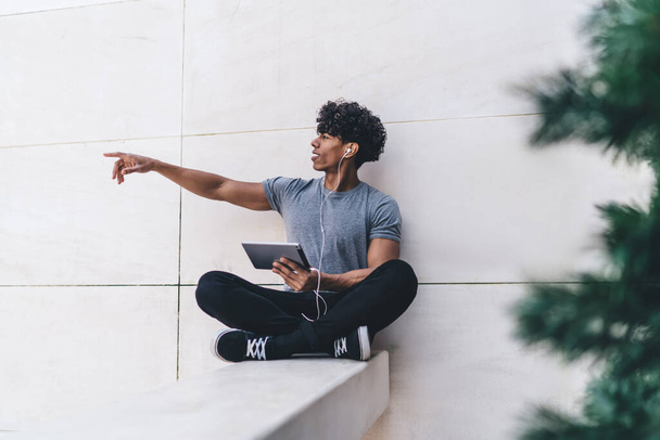 Cheerful happy handsome young African American male in casual clothes sitting legs crossed with tablet and earphones and pointing away with arm outdoors - 写真・画像
