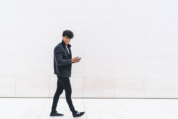 Cheerful modern adult fit male curly ethnic in black jacket and jeans surfing smartphone while walking alone on street in front of white marble wall looking at camera  - Fotó, kép