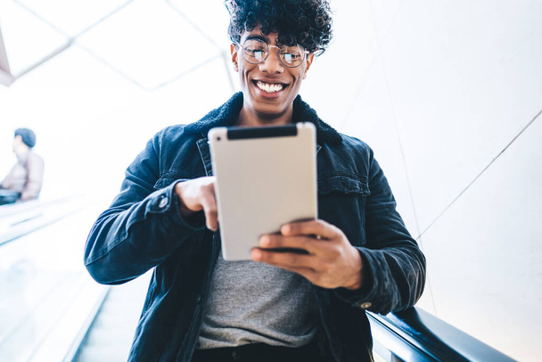Crop low angle of happy curly ethnic male in black warm jacket with round glasses riding down on escalator and using tablet on blurred background - Foto, afbeelding